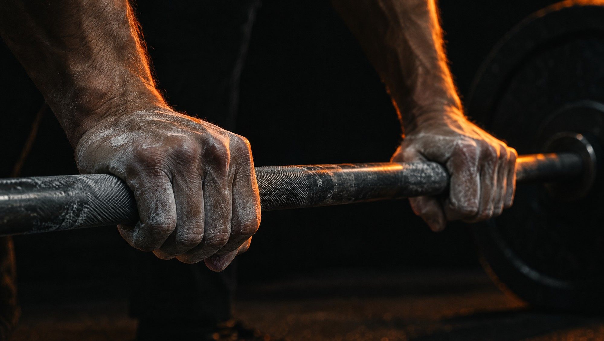 Two chalk-dusted hands gripping a knurled steel barbell in a dark gym, lit from the side in orange.