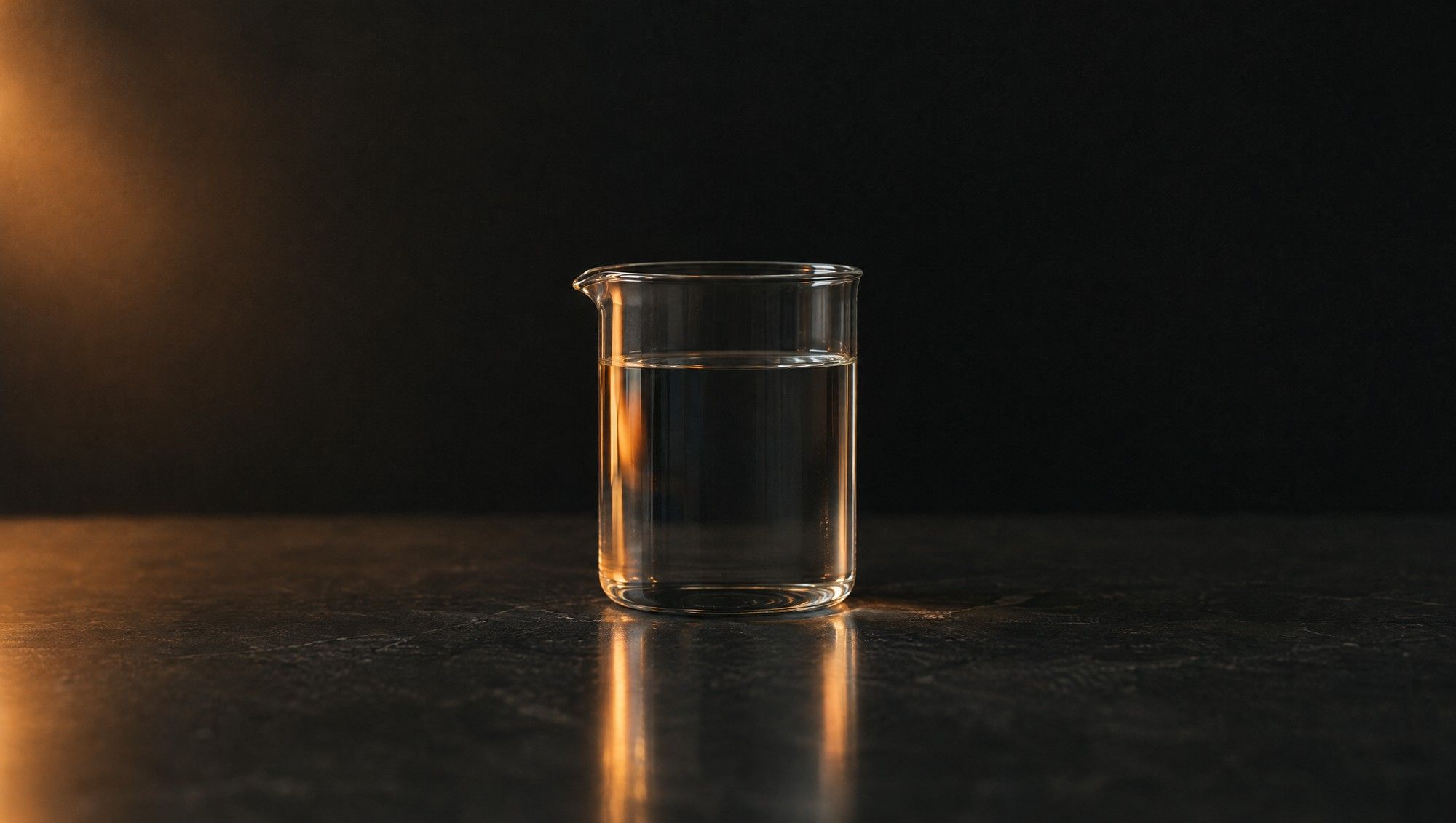 A plain glass beaker of still water on a dark stone surface, with soft orange light falling from one side.